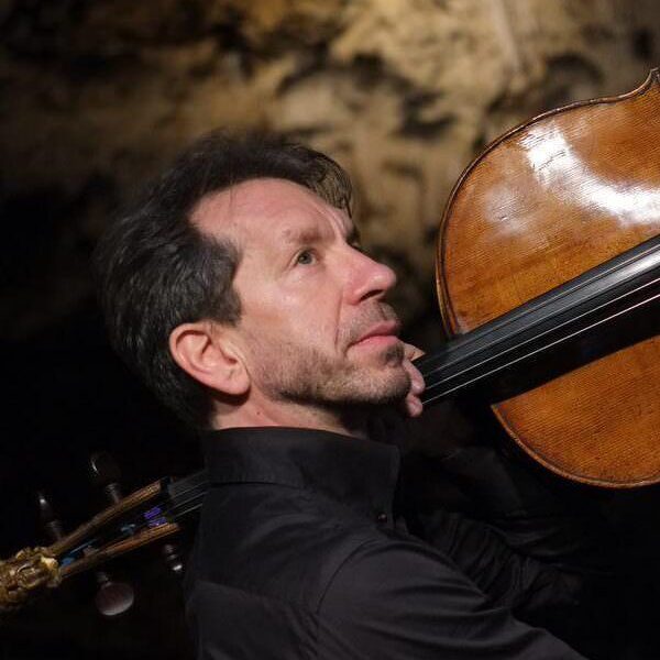 Robert Cohen holds a cello aloft over his shoulder, looking upwards.